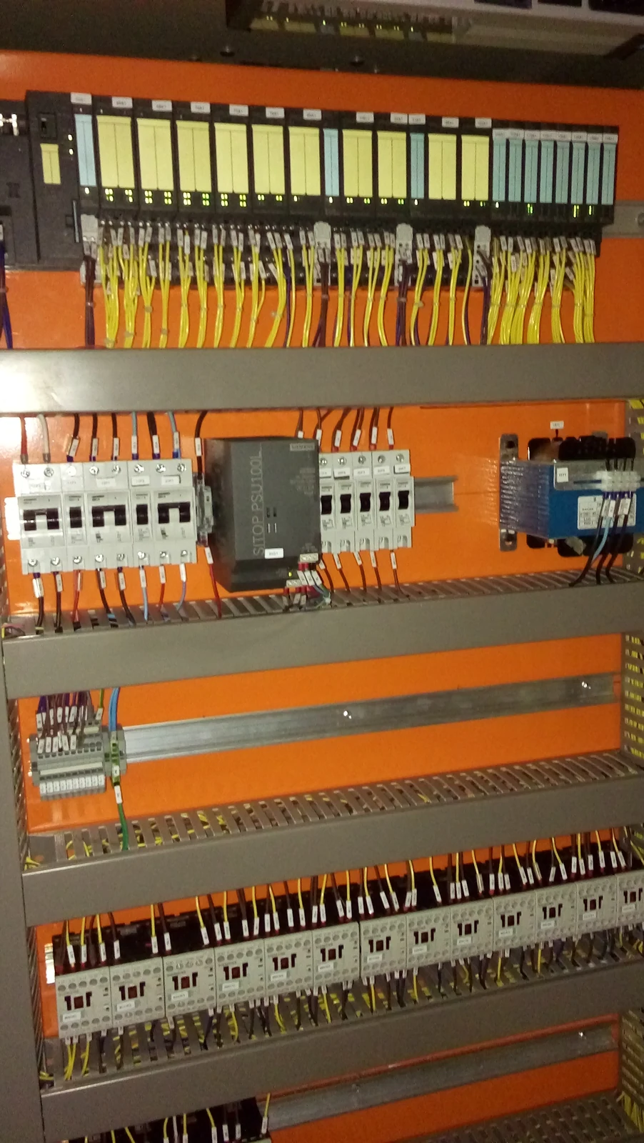 Interior de painel elétrico com componentes montados em trilhos, canaletas e condutores sobre placa laranja.